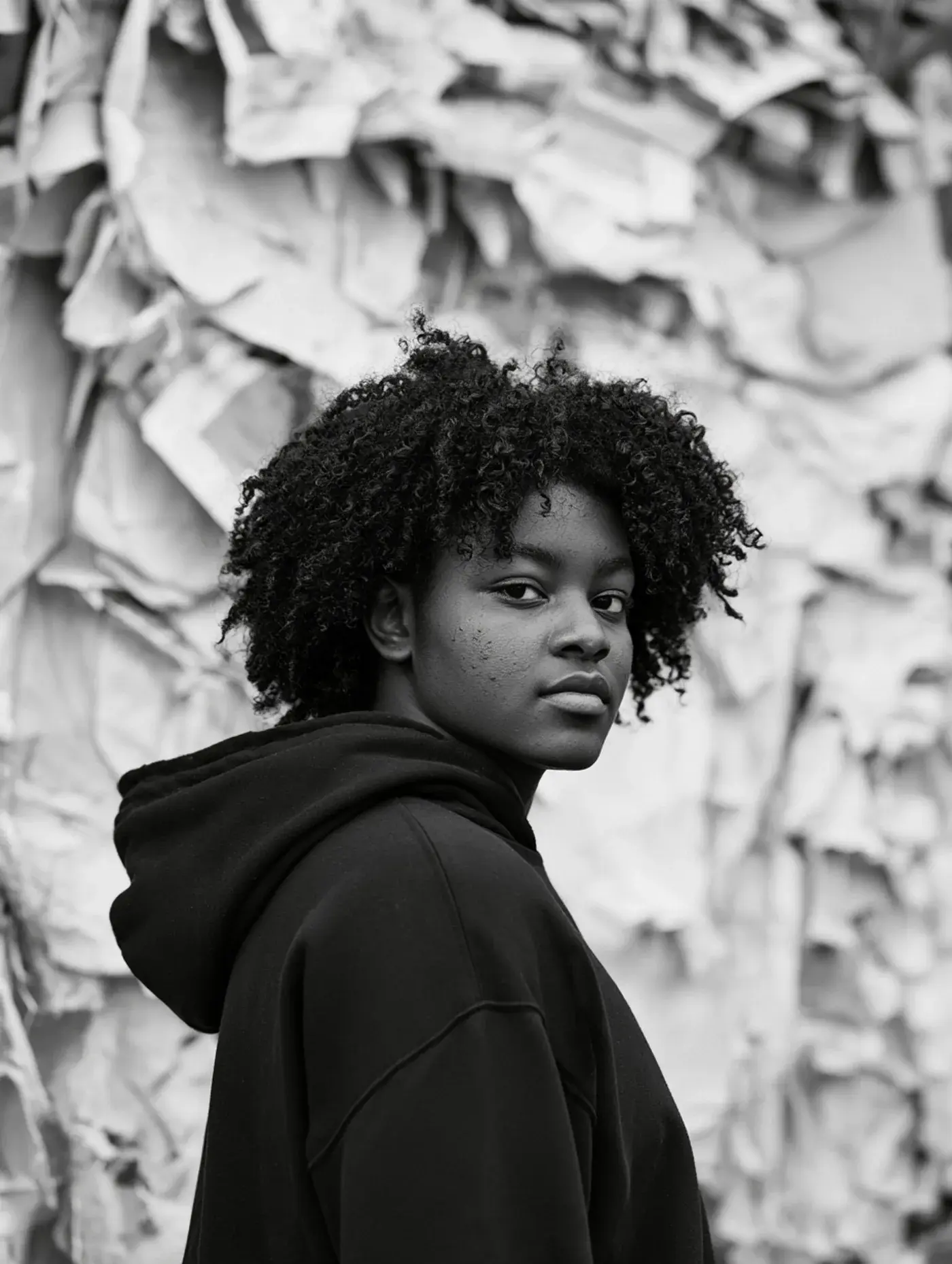 A teenage student in a black hoodie looks back over her shoulder in front of a wall of torn paper